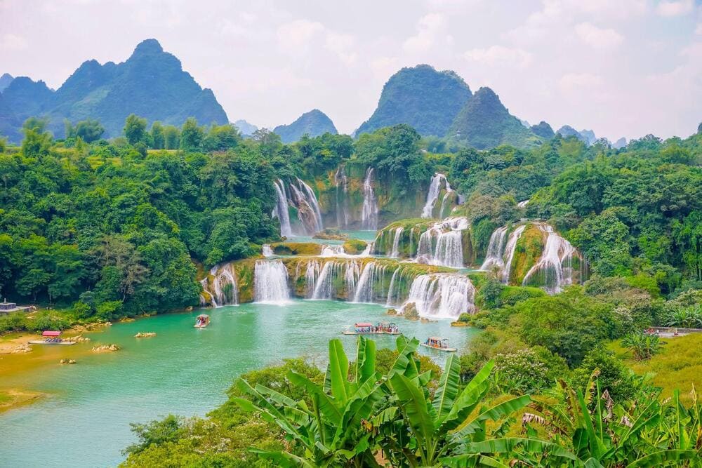 The main waterfall cascades down uneven limestone steps, creating soft white cascades that flow between lush green hills and rocky outcrops (Source: Freepik)